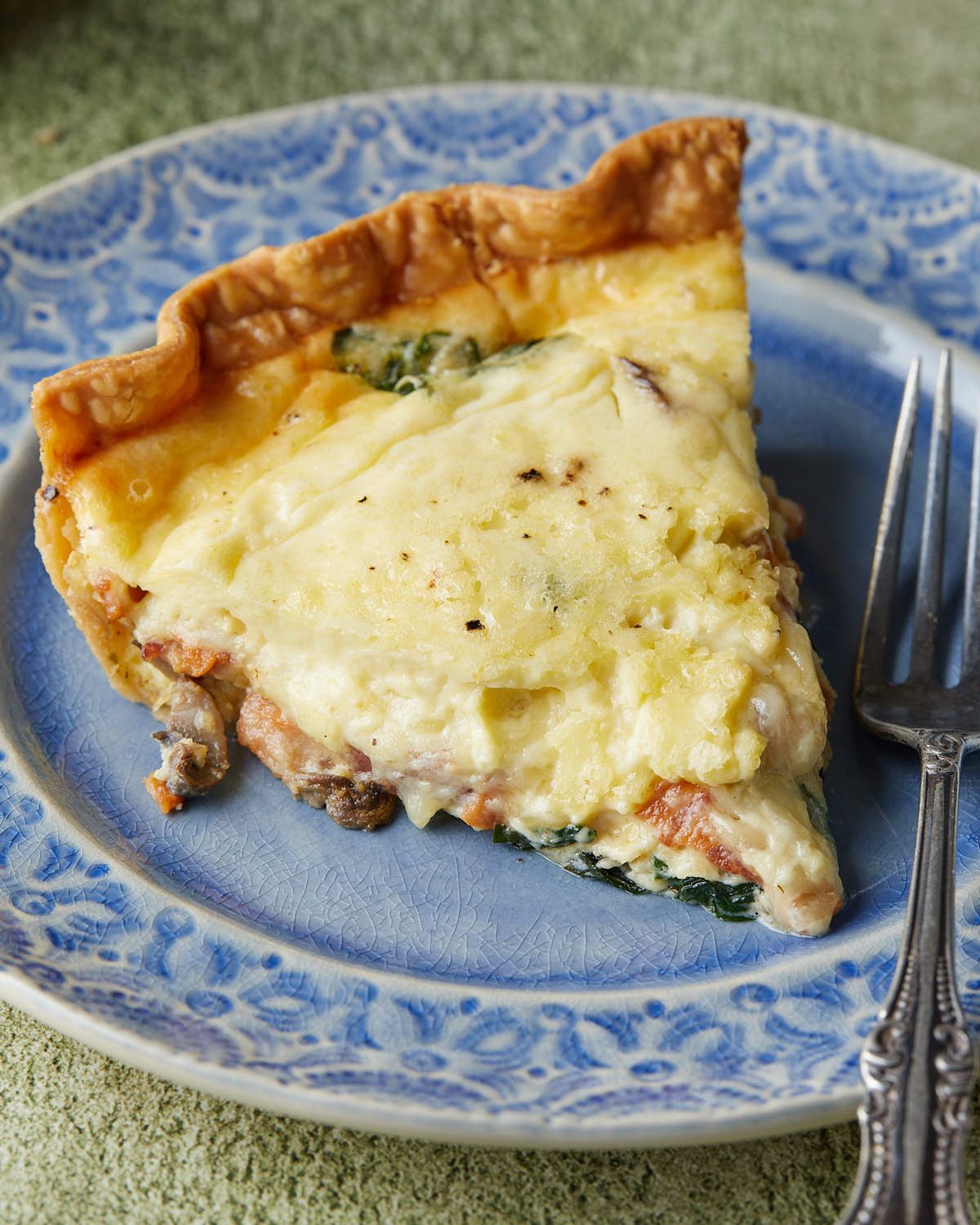 Up close image of a slice of bacon mushroom spinach quiche served on a blue plate, showing the flaky crust, creamy egg filling, crispy bacon, sautéed mushrooms, and spinach.