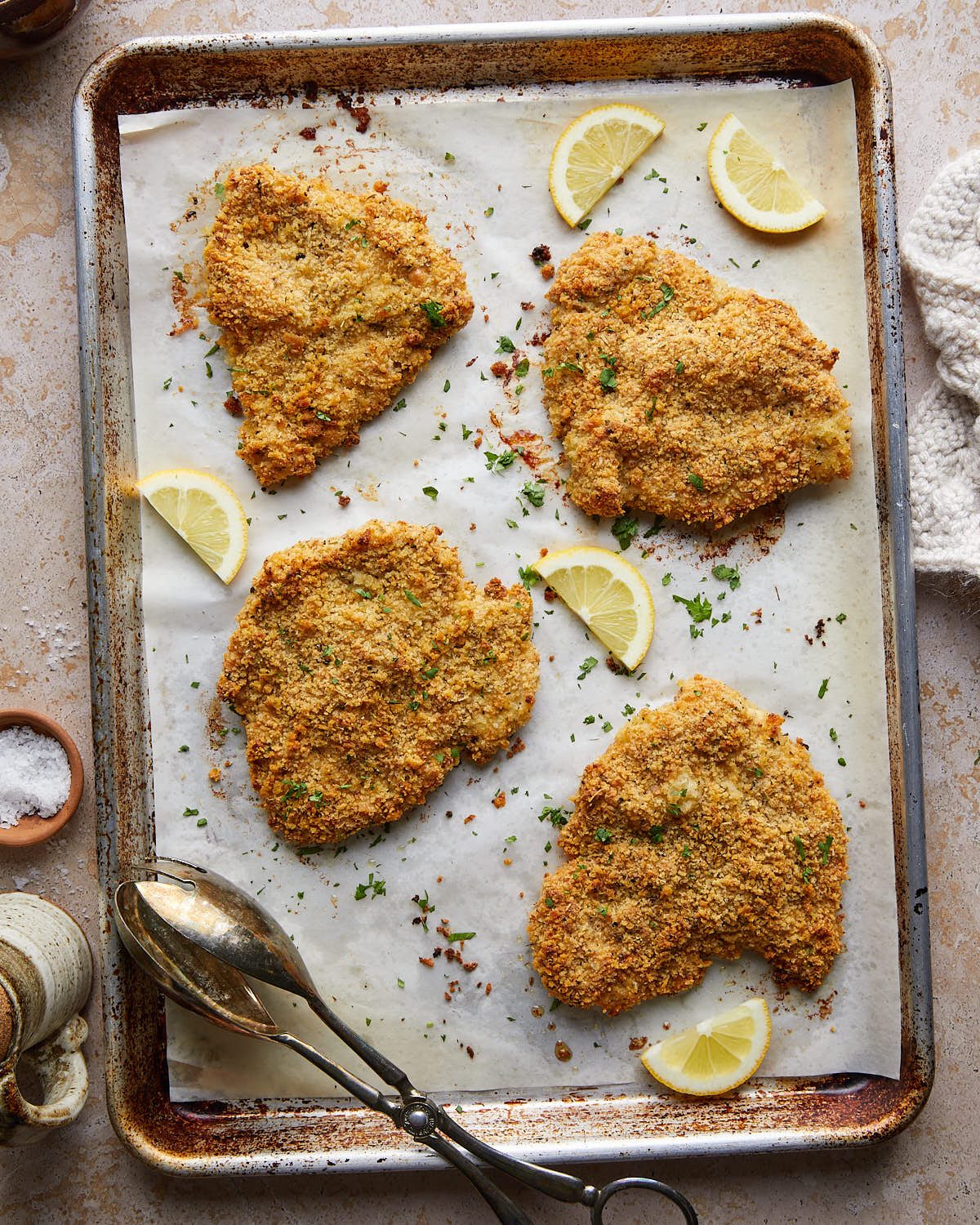 Four golden panko-Parmesan chicken cutlets on a parchment-lined sheet pan with lemon wedges and parsley.