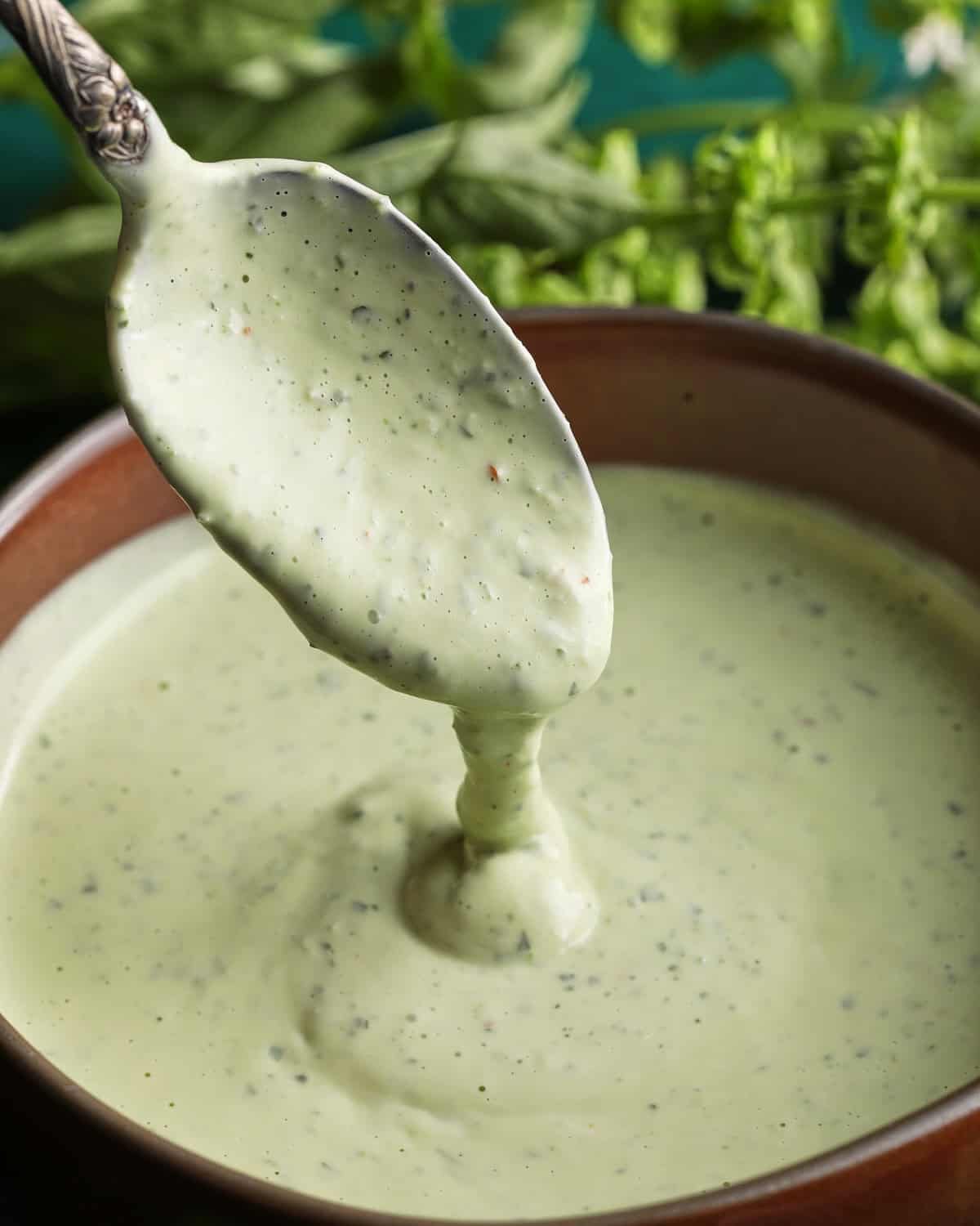 Spoon lifting creamy basil aioli from a small bowl with fresh basil in the background.