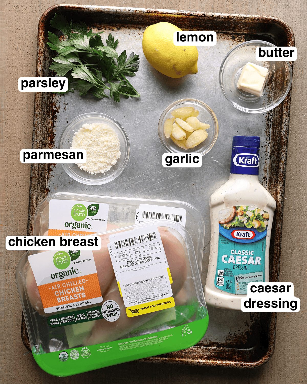 Labeled ingredients for Caesar grilled chicken arranged on a sheet pan, including chicken breast, Caesar dressing, garlic, lemon, parmesan, parsley, and butter.