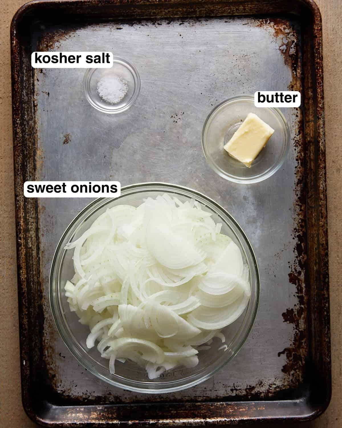 Labeled ingredients for caramelized onions arranged on a sheet pan, including sliced onions, butter, and kosher salt.