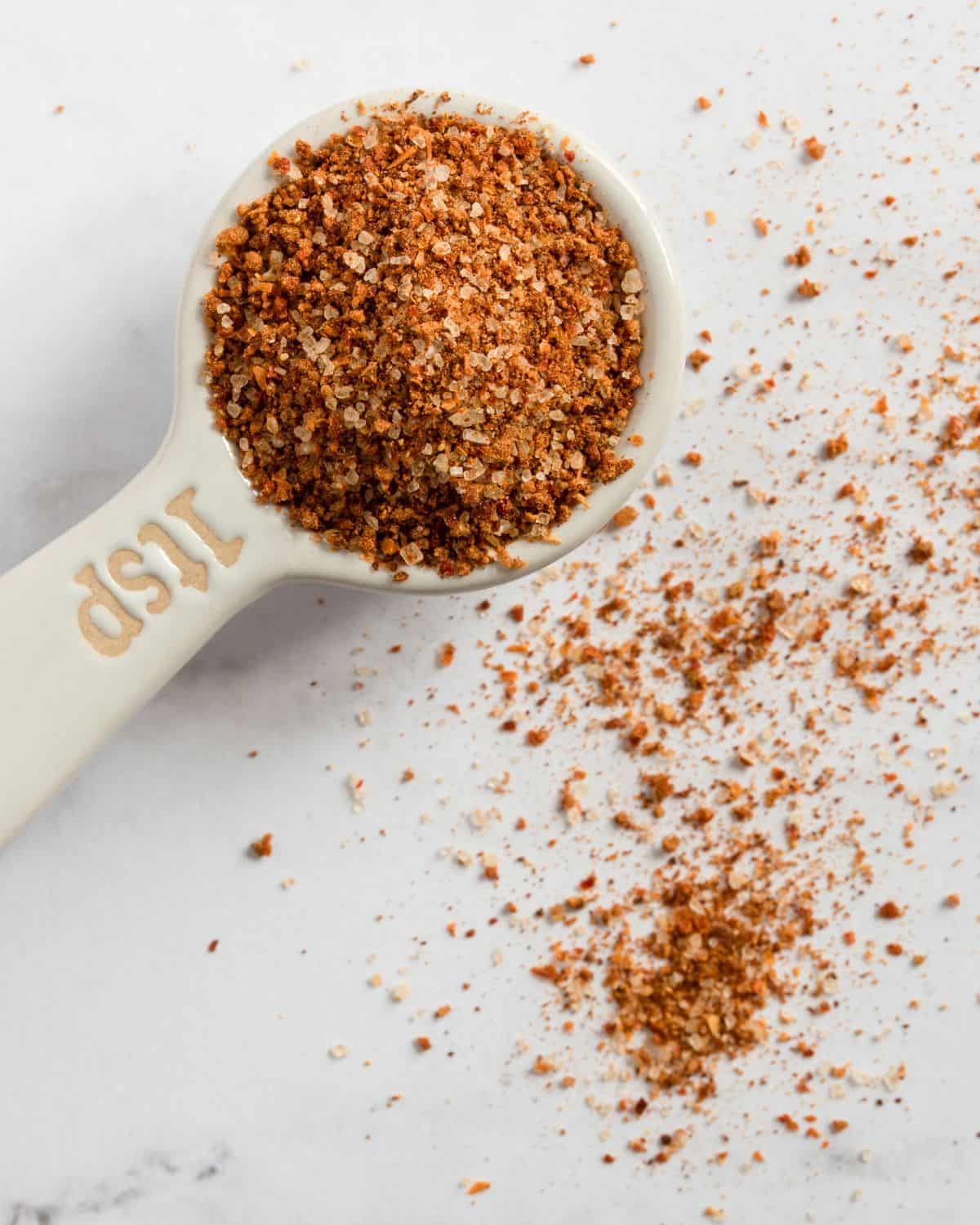 Overhead close-up of a measuring spoon filled with homemade chili seasoning recipe, showcasing a rich blend of spices with warm red and brown tones on a neutral background.