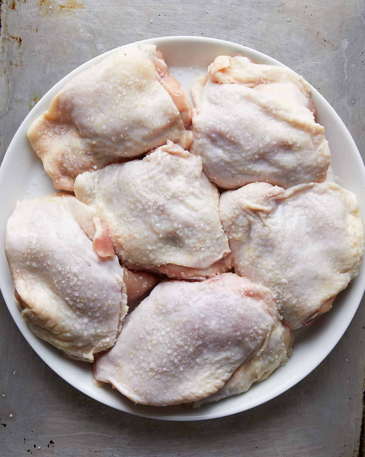 Overhead shot of dry brine chicken thighs seasoned with kosher salt on white dish.