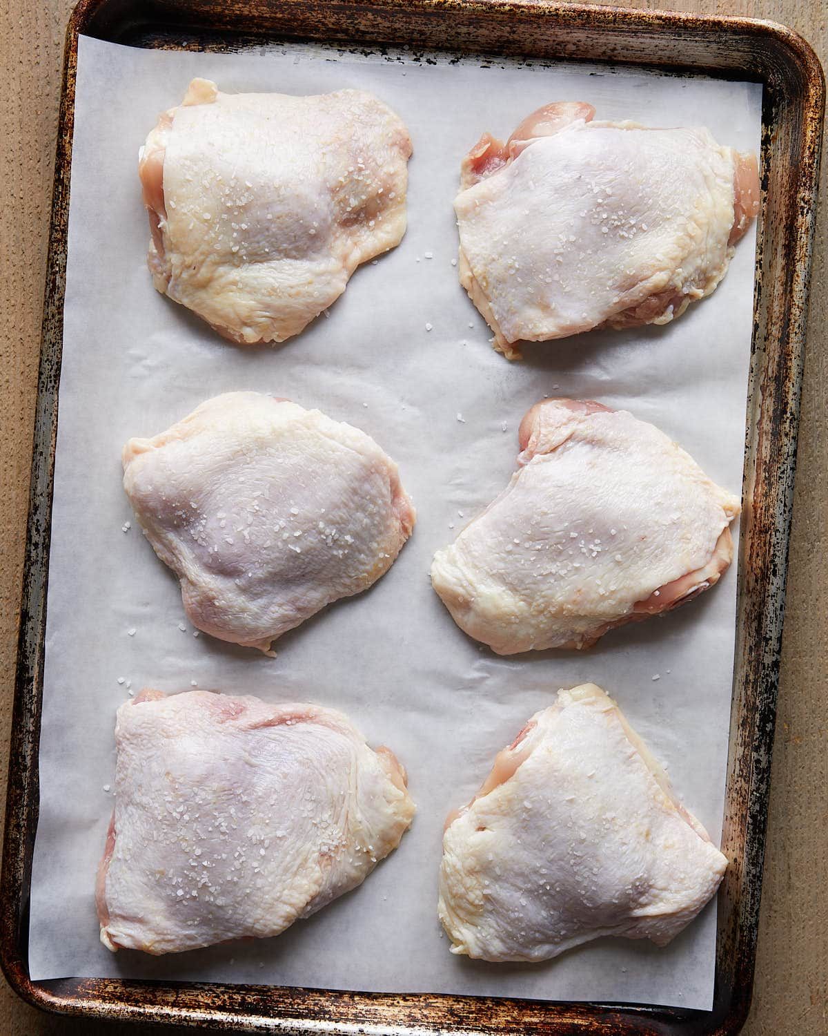 Overhead shot of dry brine chicken thighs seasoned with kosher salt, resting on a tray before cooking to enhance flavor and juiciness.