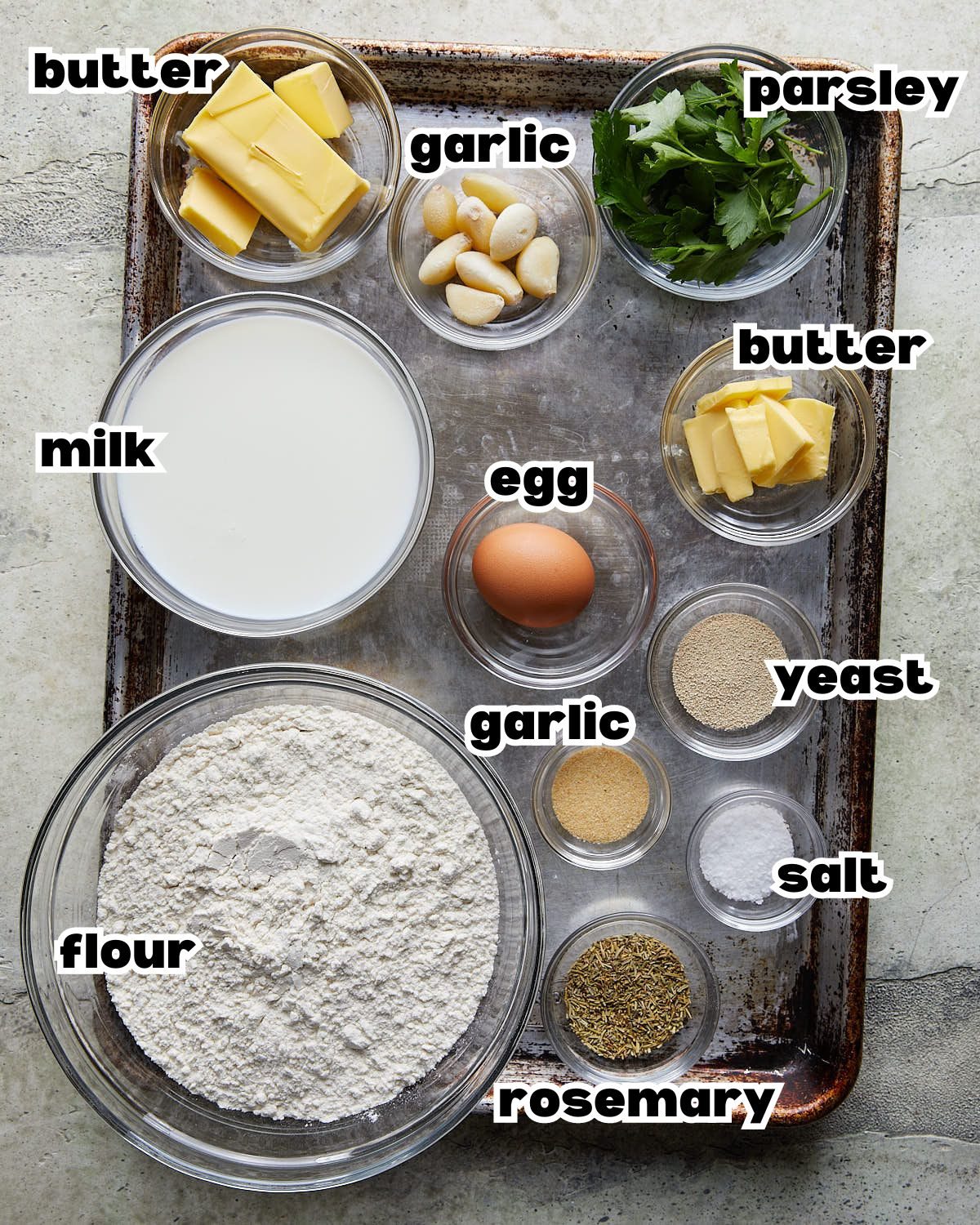 Overhead image of ingredients needed to make garlic rolls, including flour, yeast, butter, garlic, milk, herbs, and seasonings arranged in bowls.