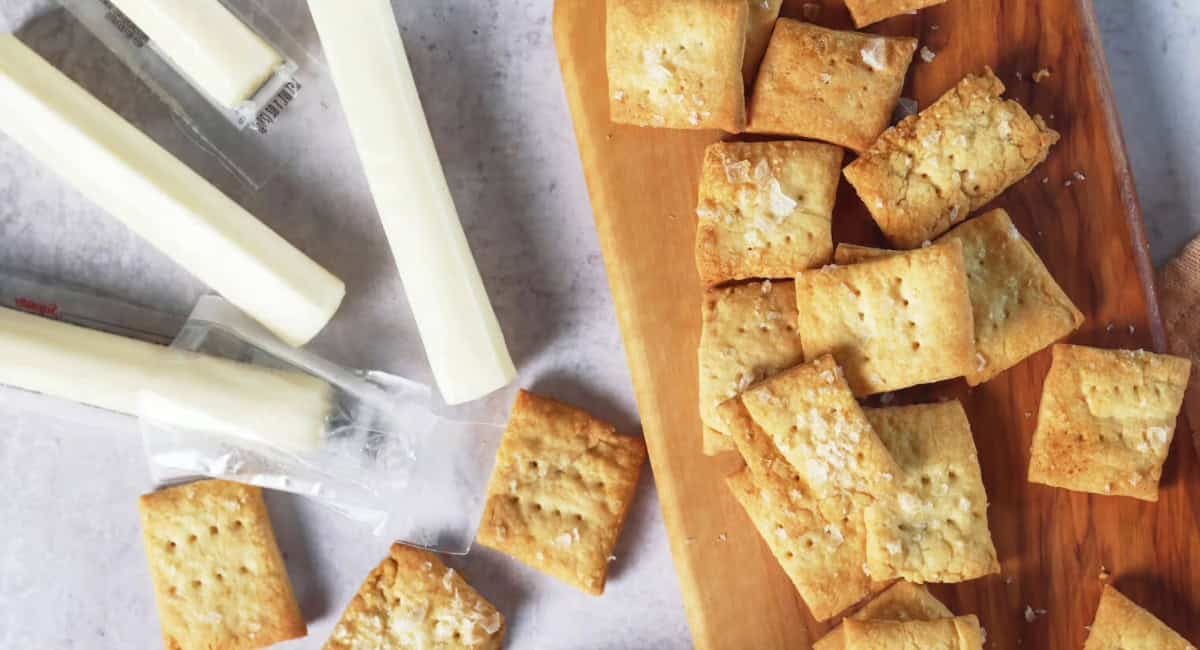 Overhead image of homemade crackers with flakey sea salt on wooden board with cheesesticks beside it.