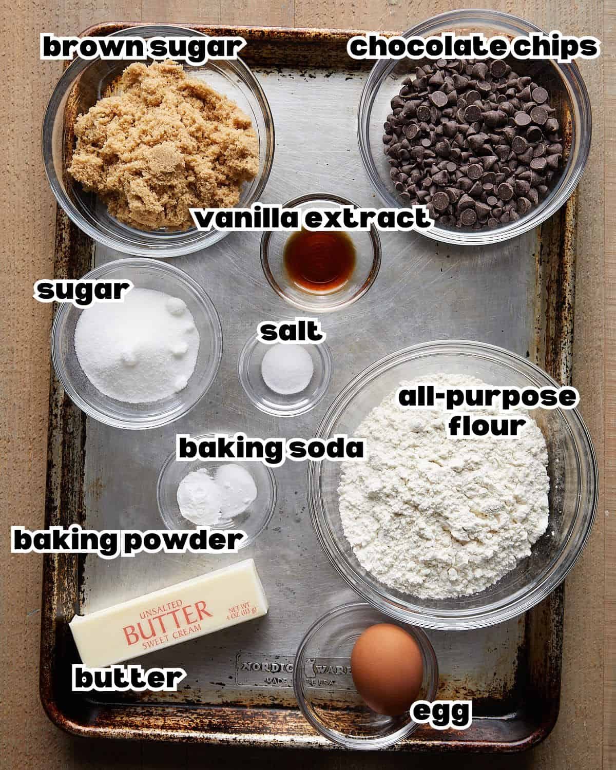 Overhead image of ingredients needed to make no chill chocolate chip cookies, including flour, butter, sugar, eggs, vanilla extract, baking soda, salt, and chocolate chips arranged on a baking sheet.