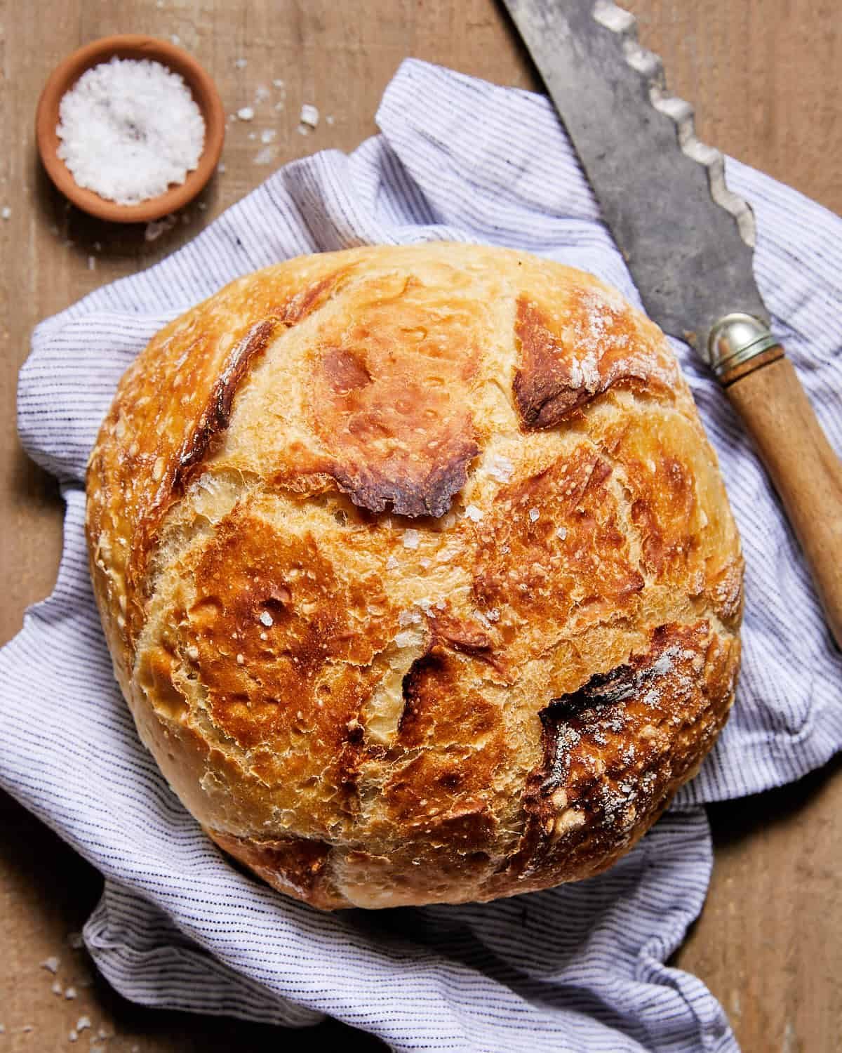 Round loaf of rustic bread with a deeply browned, craggy crust beside a bread knife on a blue-striped towel.