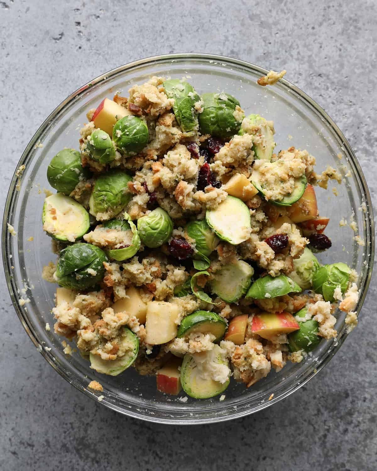Stuffing mixed with halved Brussels sprouts, diced apple, dried cranberries, and Parmesan in a glass bowl.