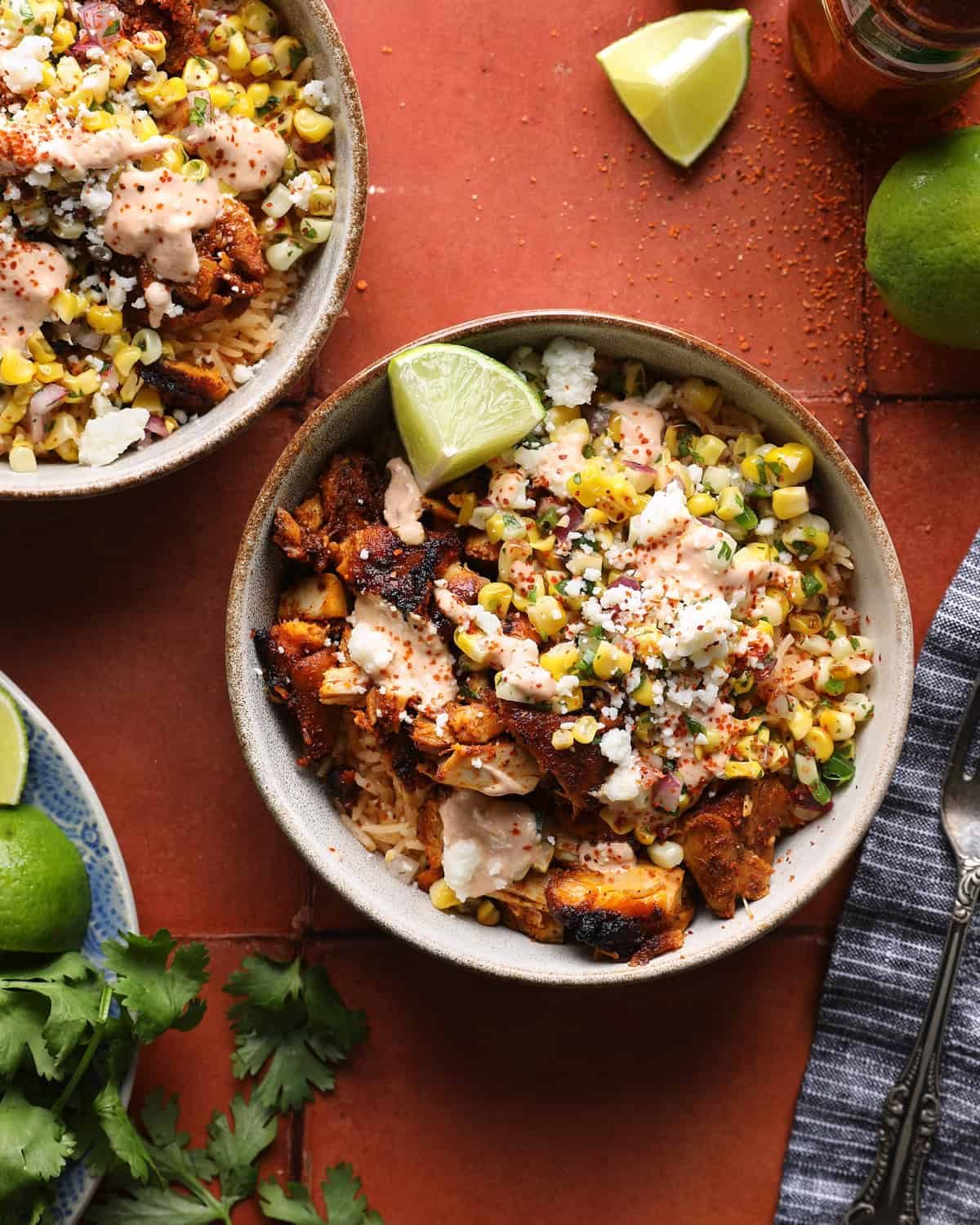 Overhead photo of a street corn chicken rice bowl with grilled chicken, corn salsa, crema, Cotija cheese, and lime wedges.