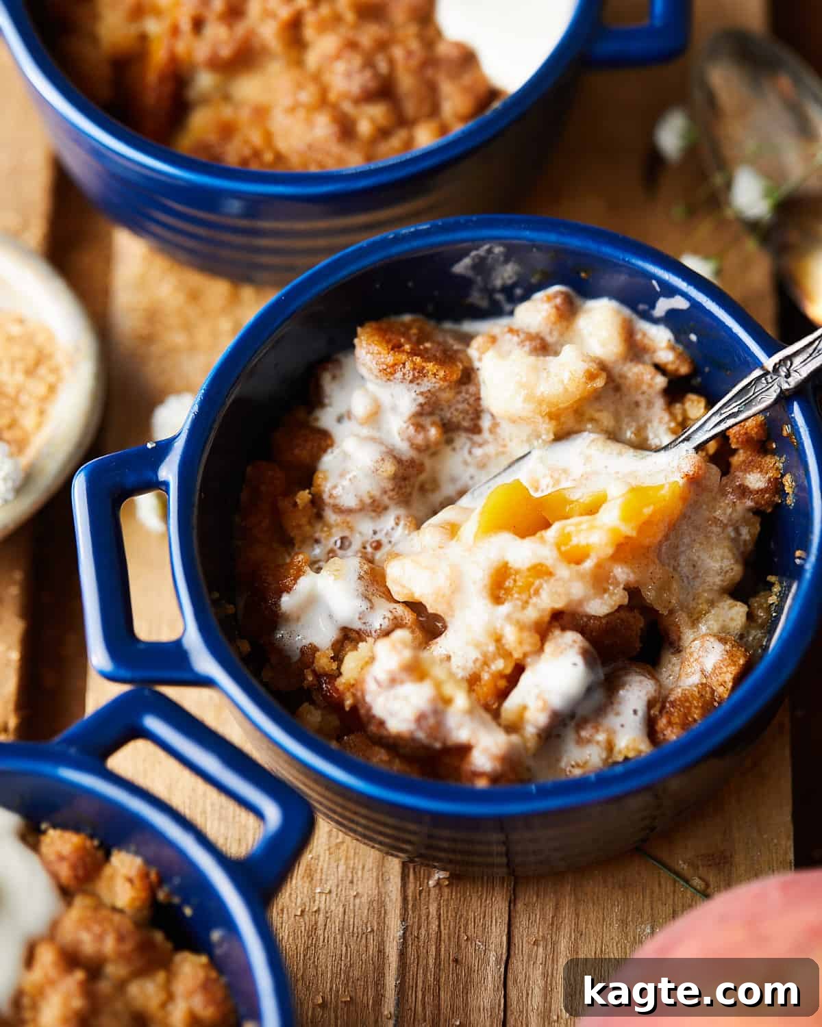 A spoon breaking into a warm serving of Air Fryer Peach Cobbler in a ramekin, revealing the soft, juicy peach filling underneath a luscious layer of melted whipped cream.
