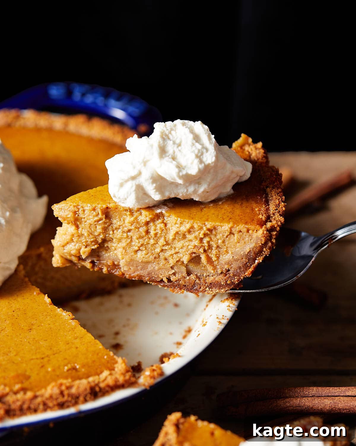 A hand gently removing a perfect slice of pumpkin pie from a blue pie dish, revealing the creamy filling and golden graham cracker crust. The slice is topped with a generous swirl of homemade whipped cream, showcasing its irresistible appeal.