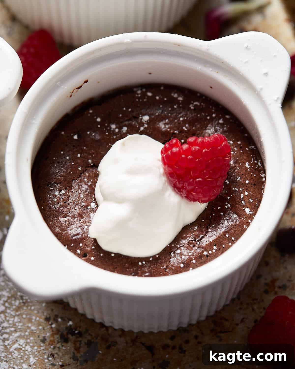 Up close image of a single mini flourless chocolate cake, warm and gooey, topped with a fresh raspberry and a dusting of powdered sugar in a white ramekin.