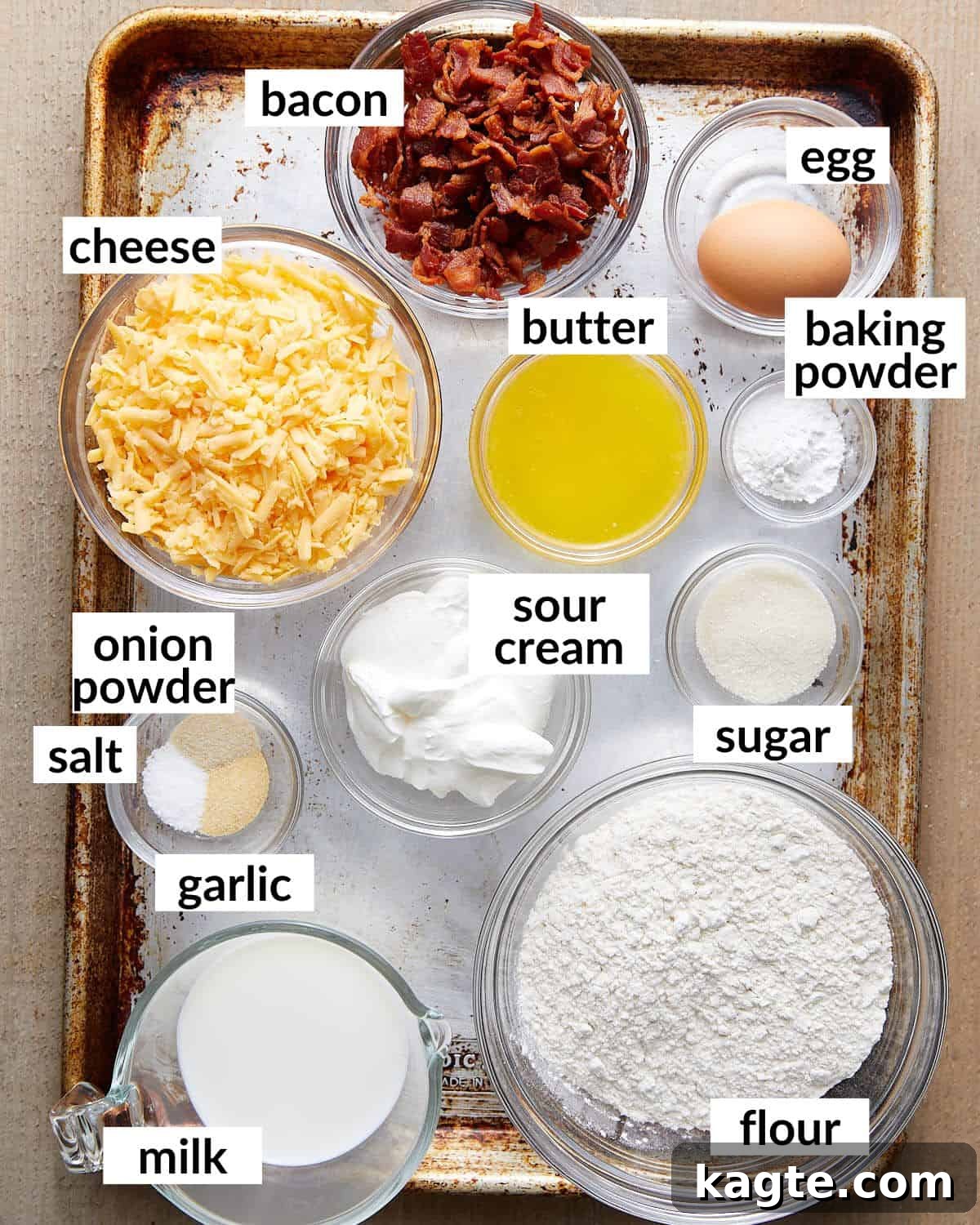 Golden Bacon Cheddar Muffins 3 Overhead image displaying all the raw ingredients neatly laid out for bacon and cheddar cheese muffins preparation: flour, sugar, baking powder, spices, milk, sour cream, butter, egg, shredded cheddar cheese, and cooked crumbled bacon.