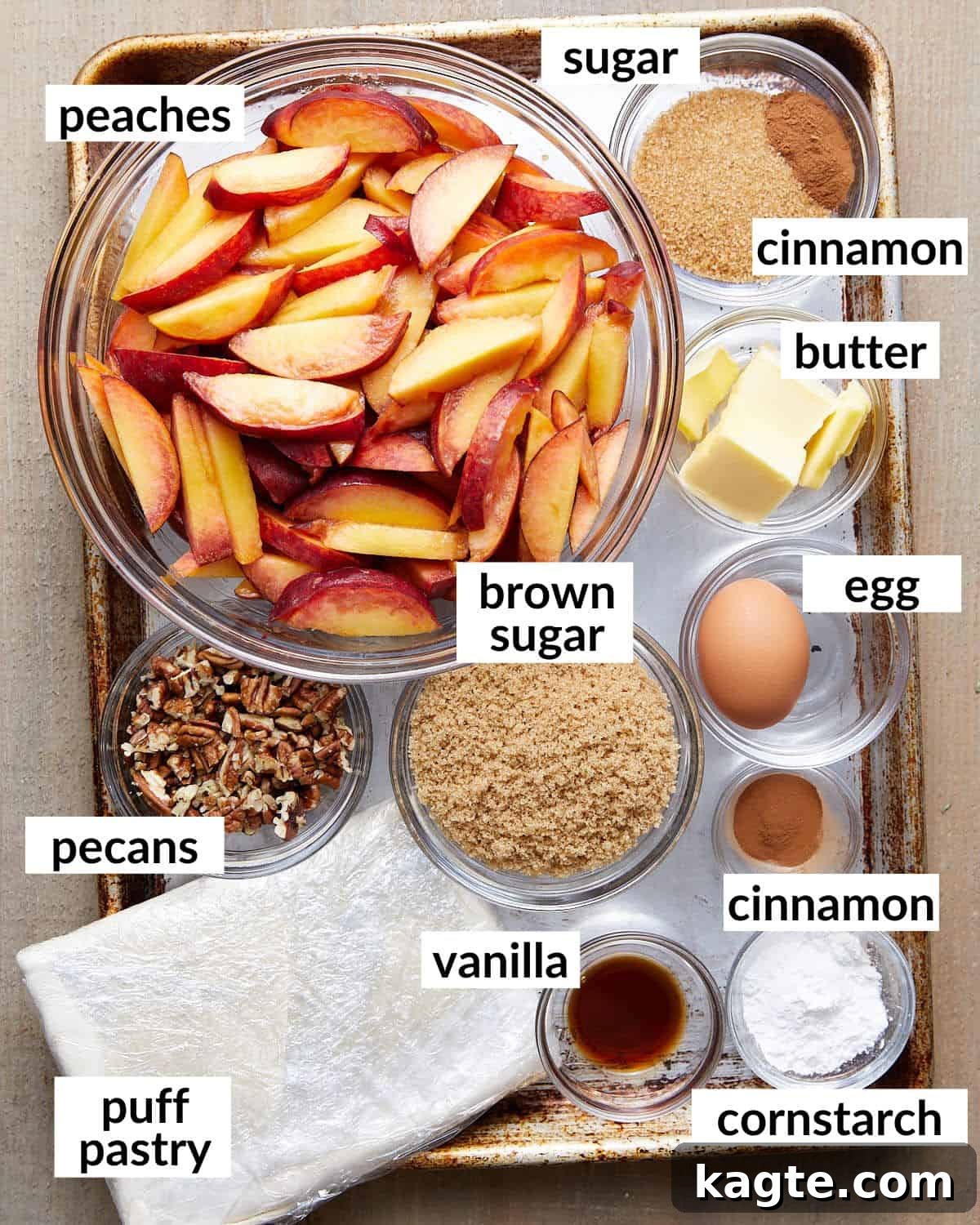 Golden Flaky Peach Cobbler 3 A selection of fresh ingredients laid out on a white surface, including sliced peaches, a block of puff pastry, brown sugar, pecans, and cinnamon, for making peach cobbler.