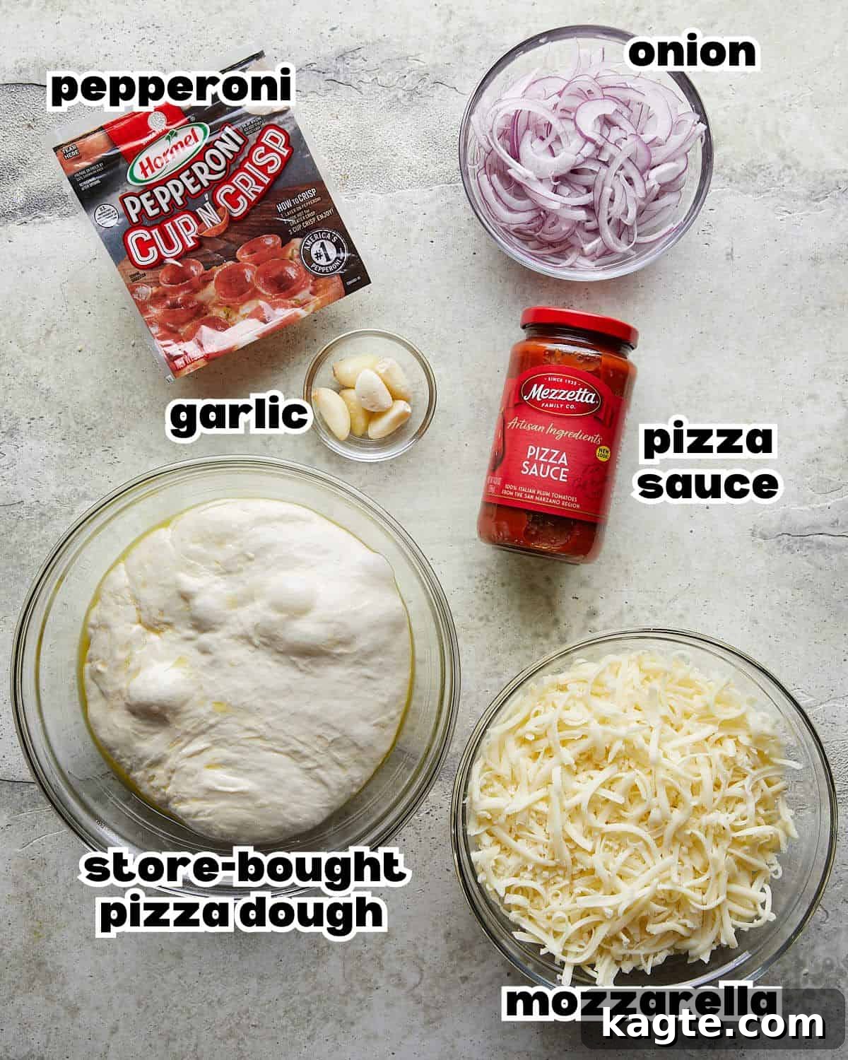 Overhead image of ingredients needed to make crispy pepperoni sheet pan pizza, including dough, garlic, sauce, cheese, onions, and pepperoni.