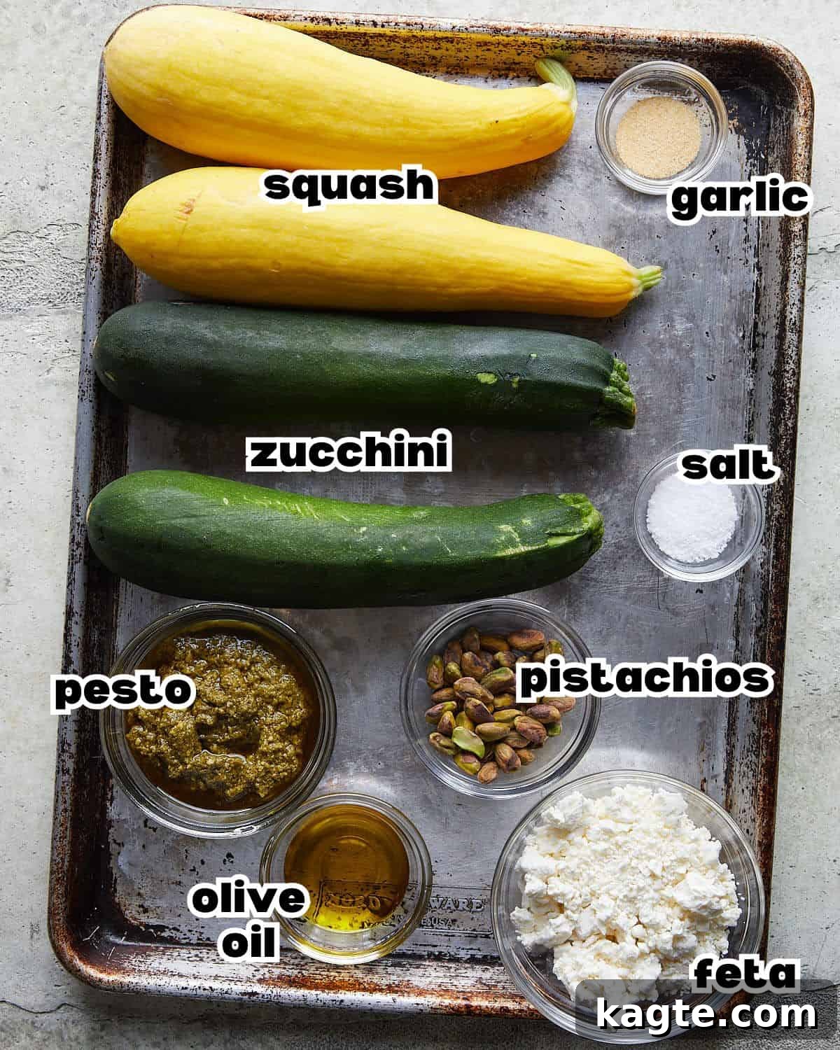Summer's Grilled Squash Medley 3 Overhead image showing all the fresh ingredients laid out for making grilled zucchini and yellow squash: fresh zucchini, yellow squash, a jar of basil pesto, olive oil, a lemon, kosher salt, granulated garlic, a bowl of pistachios, and a block of feta cheese.