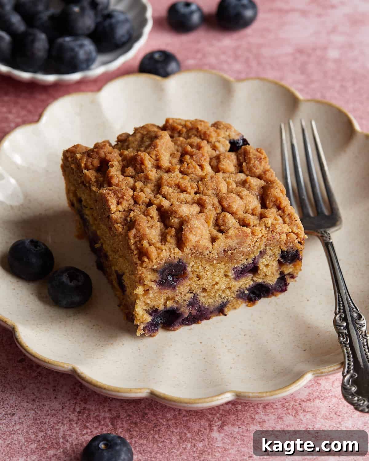 Tangy Greek Yogurt Blueberry Coffee Cake with Buttery Streusel 5 Overhead view of a slice of blueberry coffee cake served on a white dish with a fork, showing the moist interior and crumb topping.