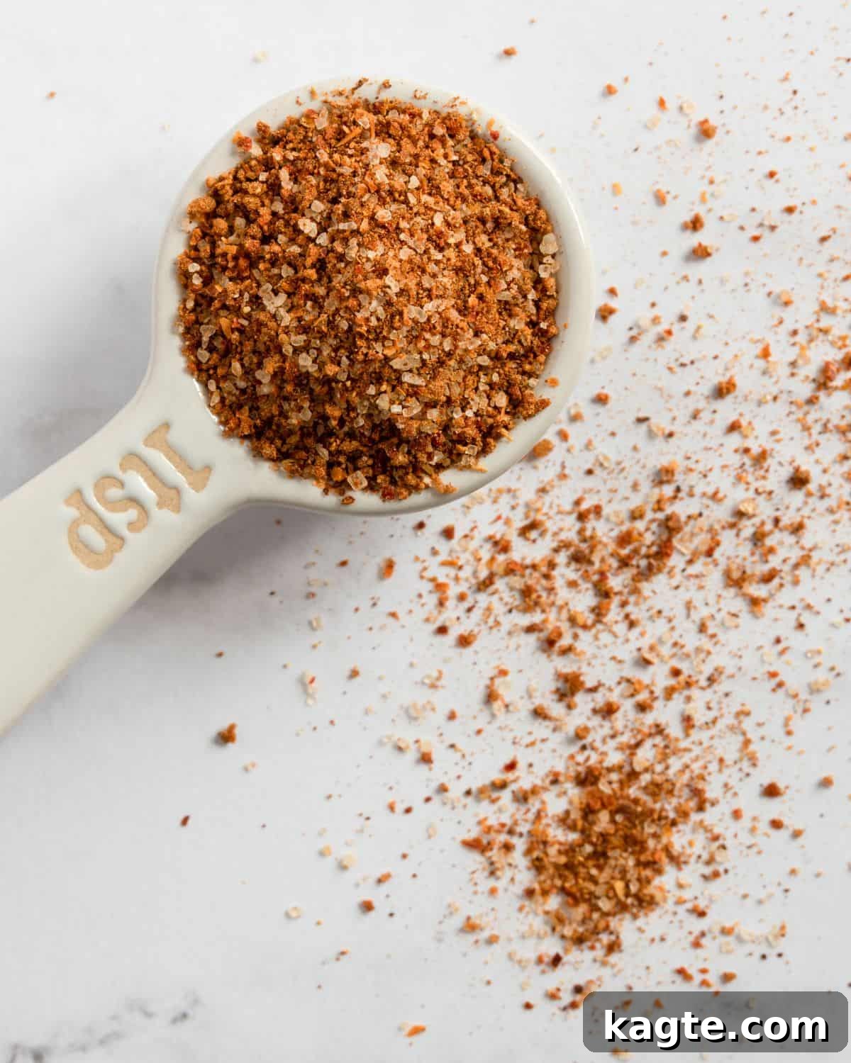 Overhead close-up of a measuring spoon filled with homemade chili seasoning recipe, showcasing a rich blend of spices with warm red and brown tones on a neutral background.