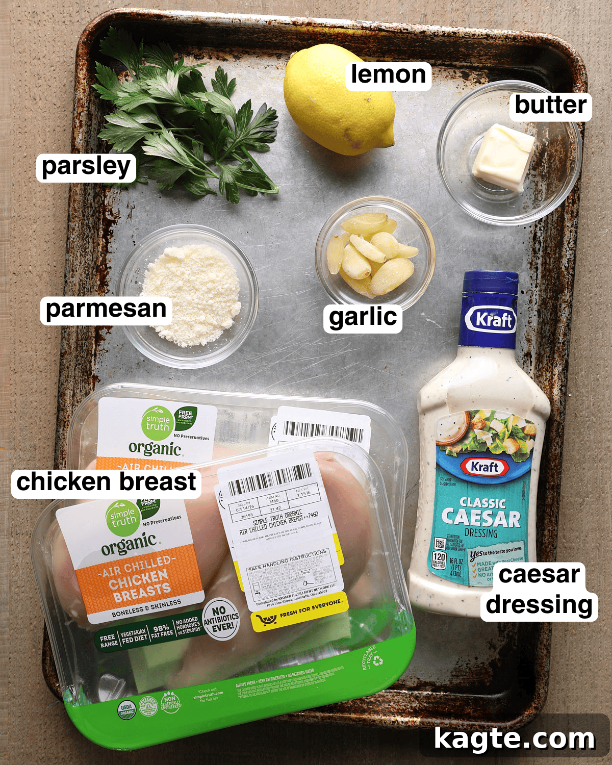 Grilled Chicken Marinated in Caesar Dressing 3 Labeled ingredients for Caesar grilled chicken arranged on a sheet pan, including chicken breast, Caesar dressing, garlic, lemon, parmesan, parsley, and butter.